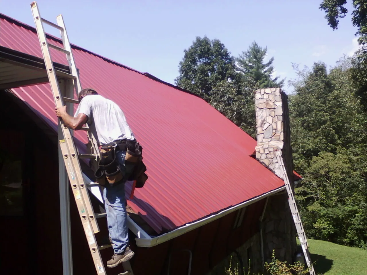 Roofer on a ladder beside a red metal roof and stone chimney — Soffit & Fascia Repair by Roofing Brothers in Buck Creek